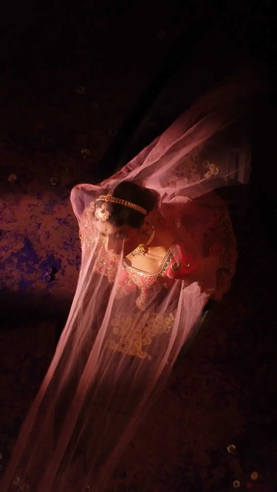 Seen from above, a rose tulle dupatta is drawn away from a bride's upturned face